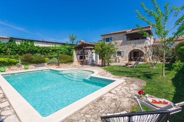 A swimming pool with stone tiles, a stone house with a tiled roof, and a grassy yard with a few trees.