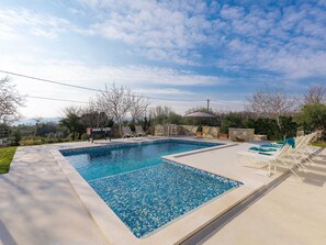 Piscina retangular com borda de mosaico, cercada por cadeiras de descanso e uma churrasqueira.