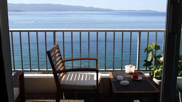 En balkon med en træstol, et lille bord med et lys og en skål, samt udsigt til havet og fjerne bakker.