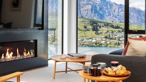 A cozy living room with a fireplace, a coffee table with books, a cup, and pastries, and a view of mountains and a lake.