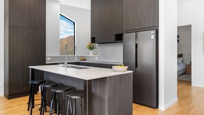 A modern kitchen with a central island, stainless steel appliances, and a view of a mountainous landscape through a window. - A Twist on Olivers | Luxury | Lake and Mountain Views (Queenstown)