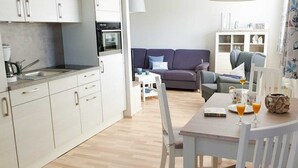 Una cocina compacta con armarios blancos y un horno empotrado. Se observa un comedor con una mesa y sillas, con un sofá y un mueble de fondo.