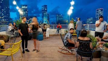 A rooftop bar with a city skyline view, wooden deck, and various seating options.