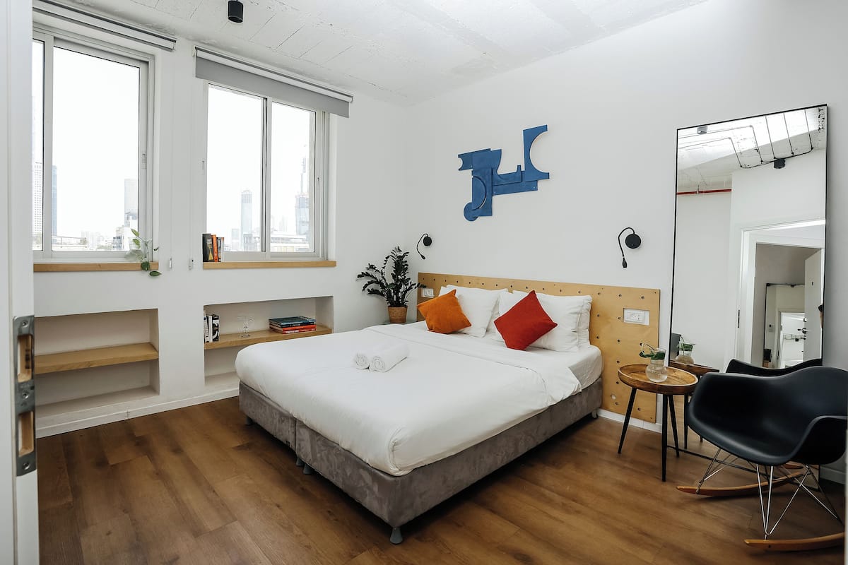 Quarto de hotel moderno com uma cama grande, uma mesinha de cabeceira, uma cadeira e um espelho amplo.