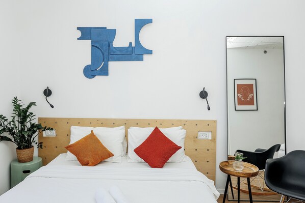 Une chambre d’hôtel bien rangée, avec un lit, une petite table ronde en bois, une chaise noire, une plante en pot et un grand miroir au mur.
