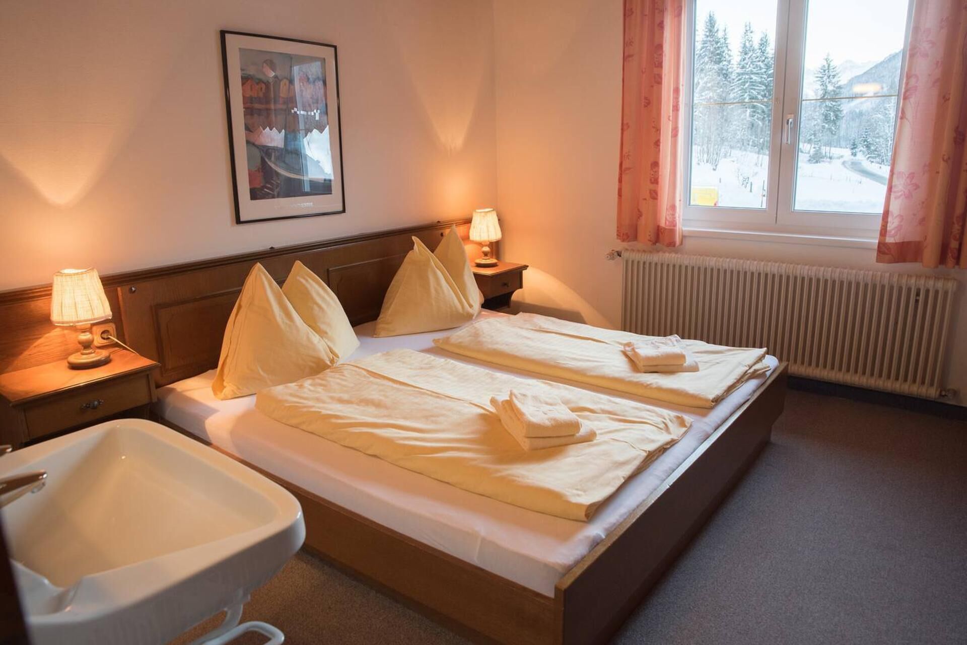 Ein Hotelzimmer mit zwei Betten, einem Badezimmer, einem Heizkörper und einem Fenster mit Blick auf schneebedeckte Bäume.