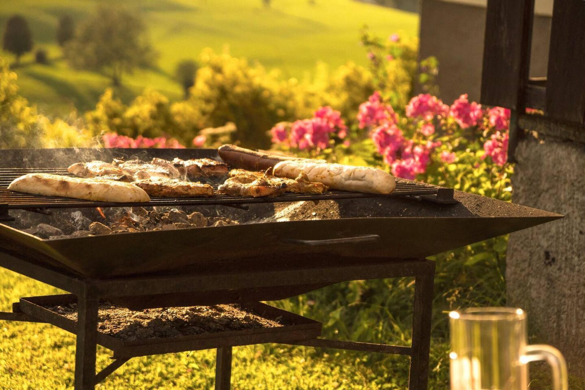 A barbecue grill with food being cooked outdoors, surrounded by greenery and flowers.