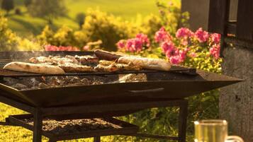 A barbecue grill with food being cooked outdoors, surrounded by greenery and flowers.