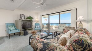 A spacious living room with a large TV, comfortable seating, a ceiling fan, and a view of the beach through large windows.