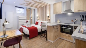 A modern hotel room with a bed, bedside table, lamp, and a kitchenette with a stove and sink. - Les Studios de la faisanderie 1 (Chantilly)