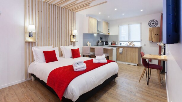 A modern hotel room with a bed, a desk, a chair, a kitchen area, and a clock on the wall. - Les Studios de la faisanderie 1 (Chantilly)