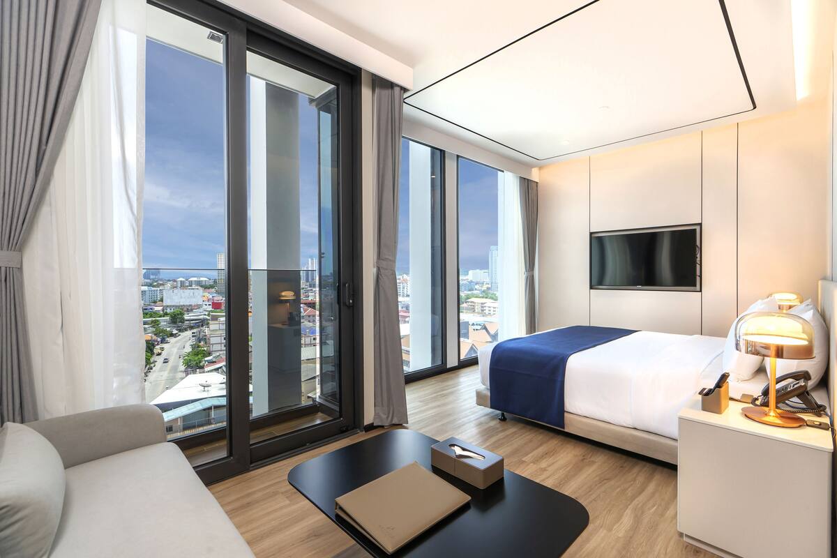 A modern hotel room with a large window, a bed, a sofa, a desk, and a TV.