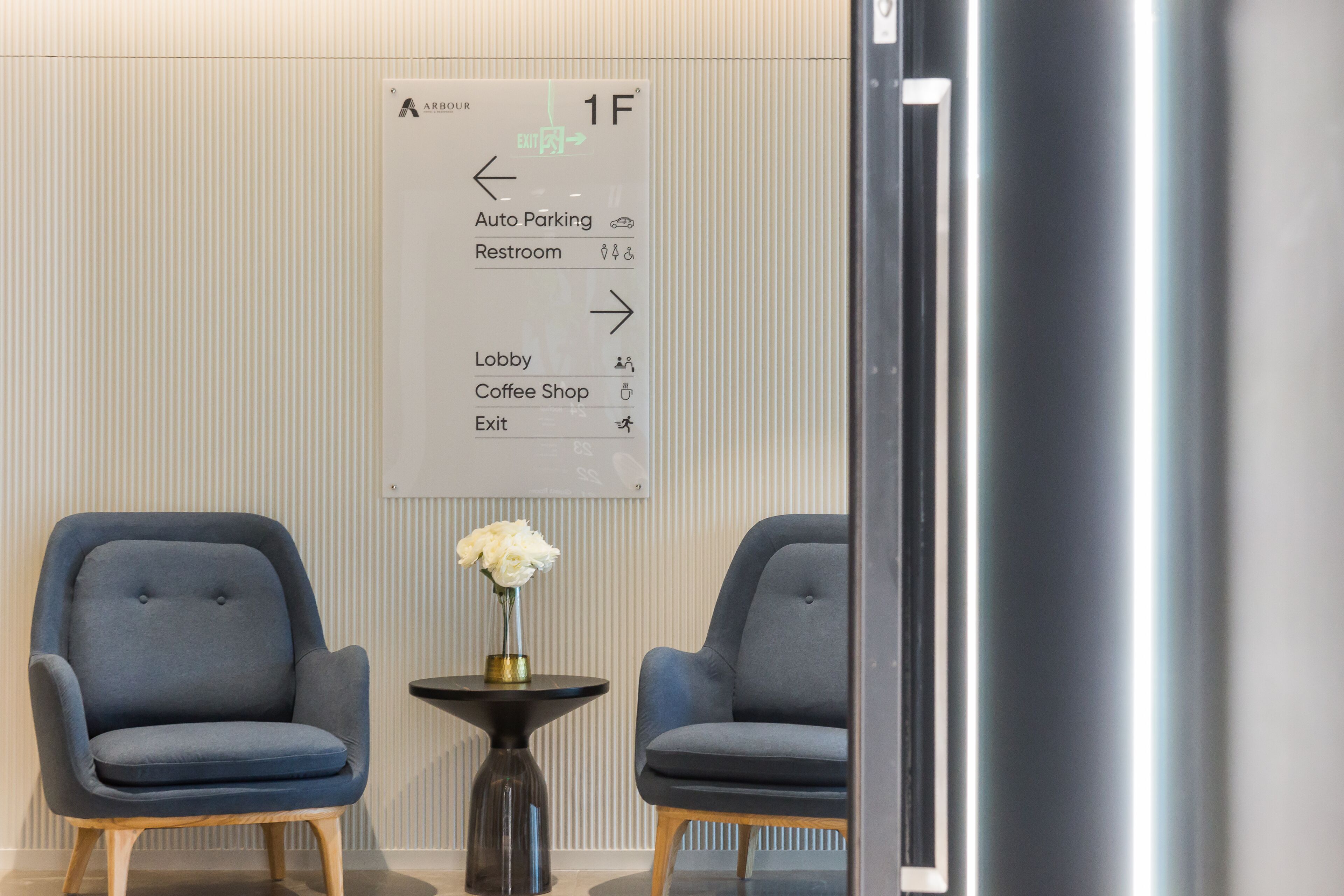 A modern lobby with directional signage, two blue armchairs, a small table with a vase of flowers, and a metallic column.