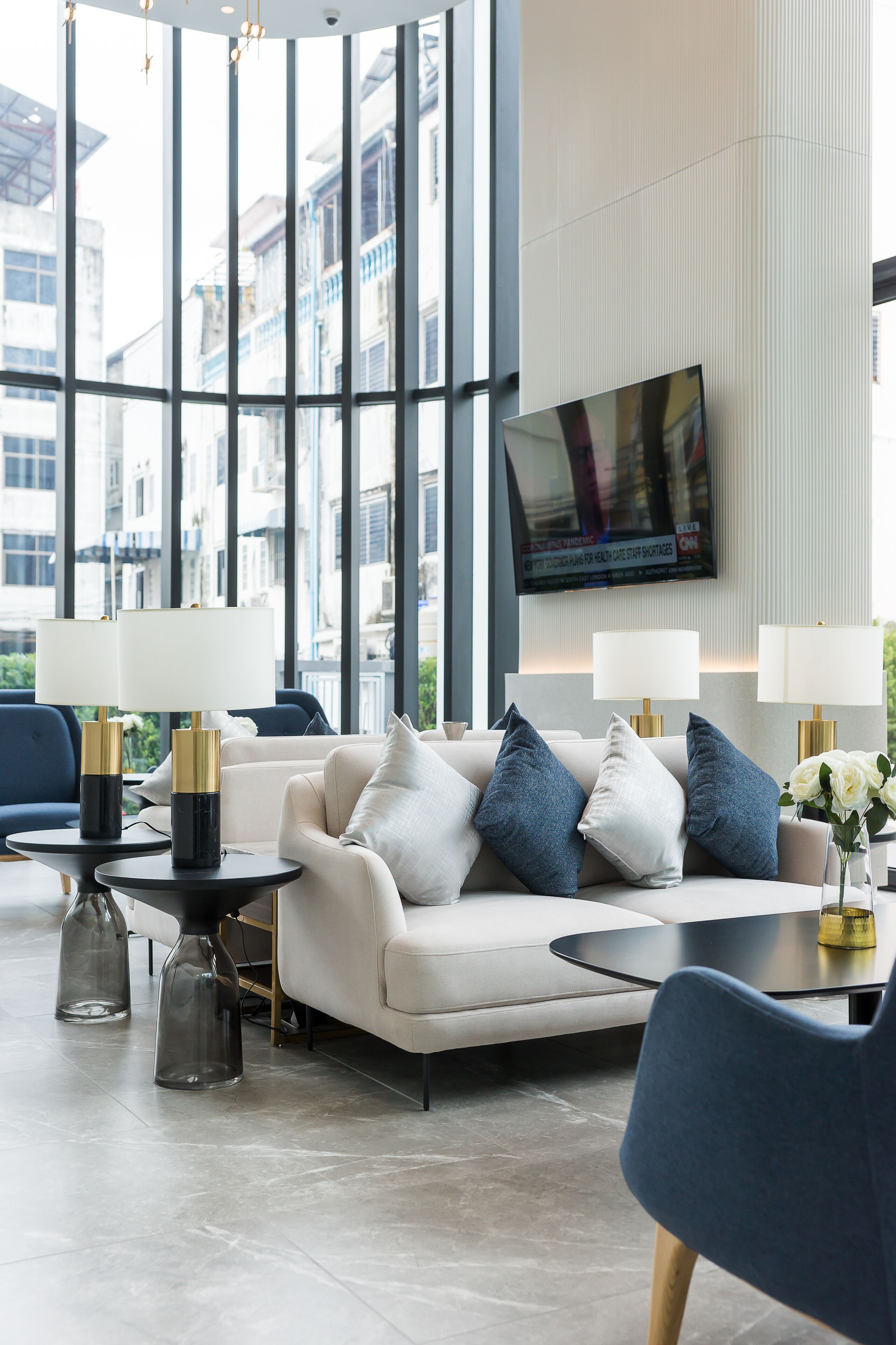 A modern hotel lobby with a large window, a flat-screen TV mounted on the wall, a sofa with cushions, and two side tables with lamps.