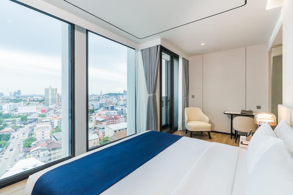 A hotel room with a large window offering a city view, a bed with white and blue bedding, a desk with a chair, and a small table with a lamp.