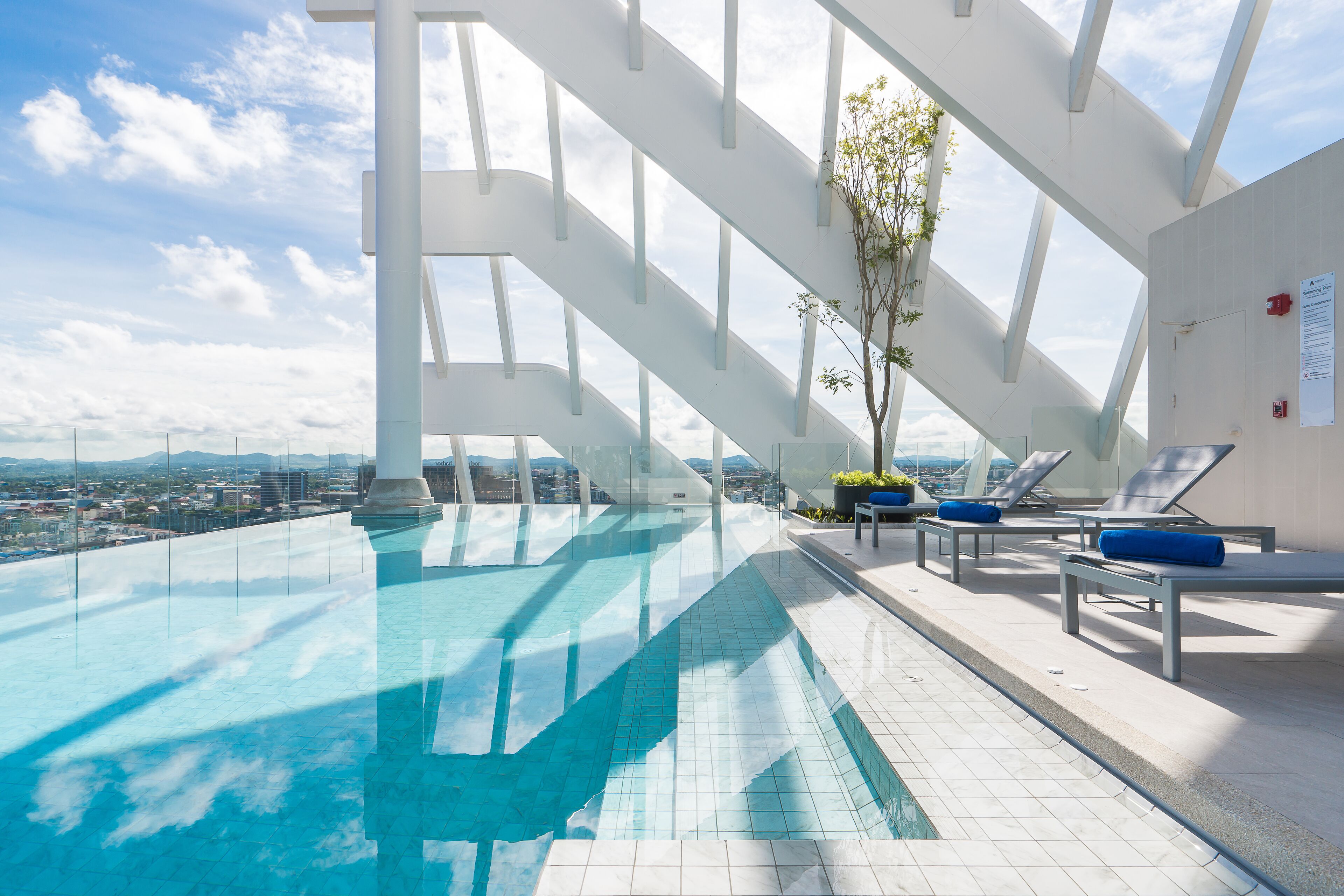 An infinity pool with a clear view of the cityscape, white structural beams, and lounge chairs with blue cushions.