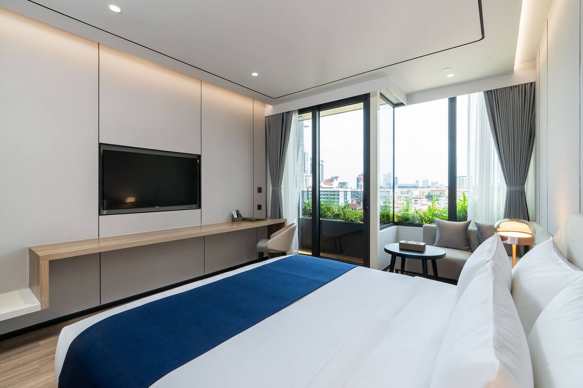 A modern hotel room with a large bed, a flat-screen TV, a sofa, a small table, and a balcony with a city view.