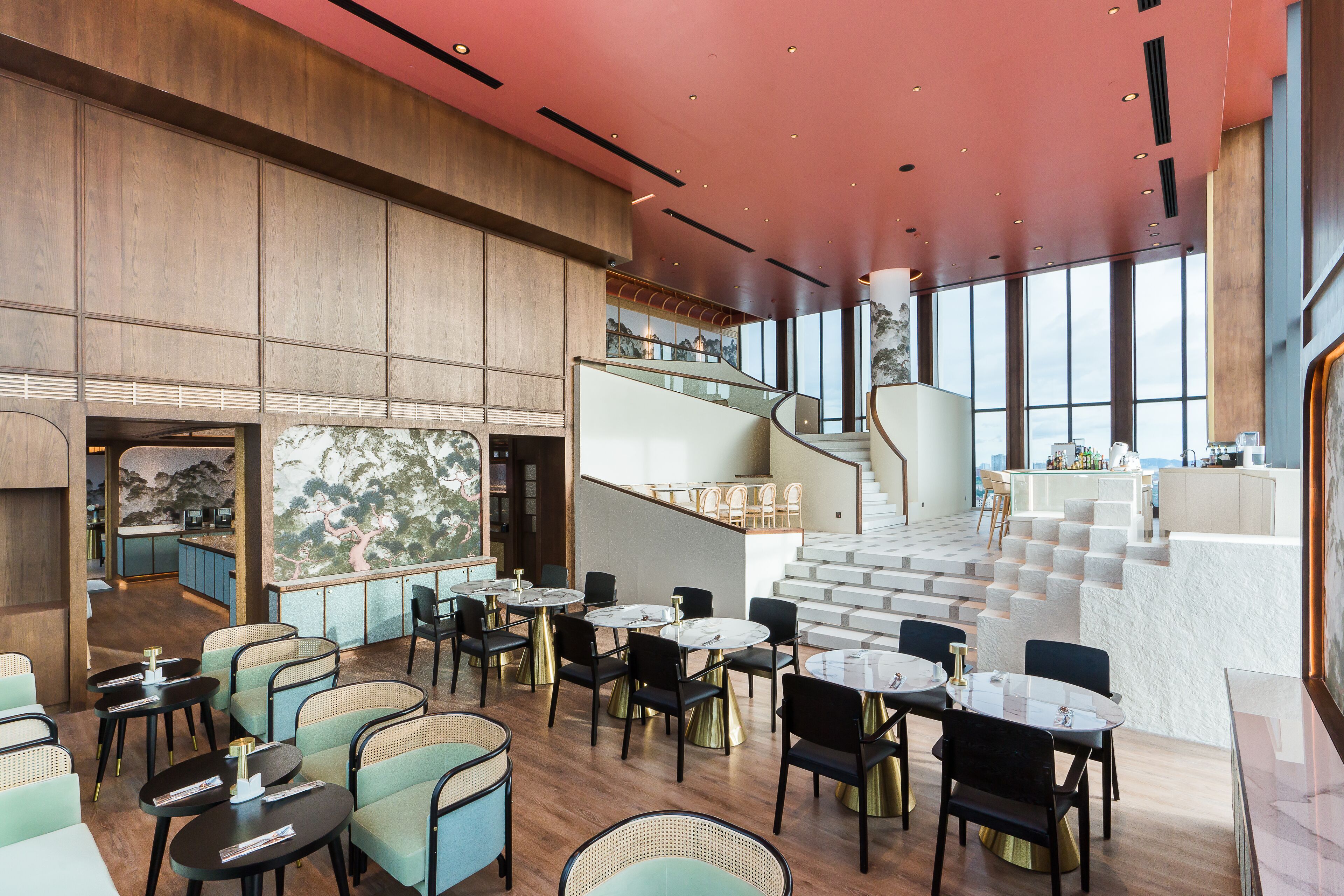 A spacious restaurant with high ceilings, a staircase leading to an upper level, and a view of the cityscape through large windows.