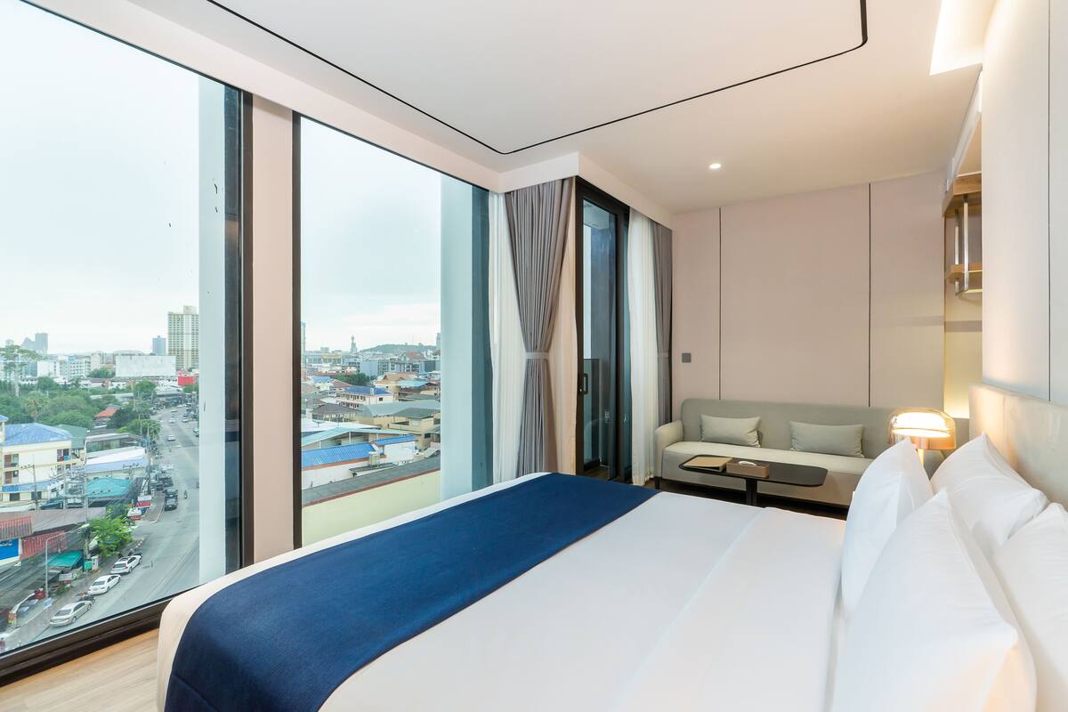 A modern hotel room with a large bed, a sofa, a small table, and a city view through large windows.