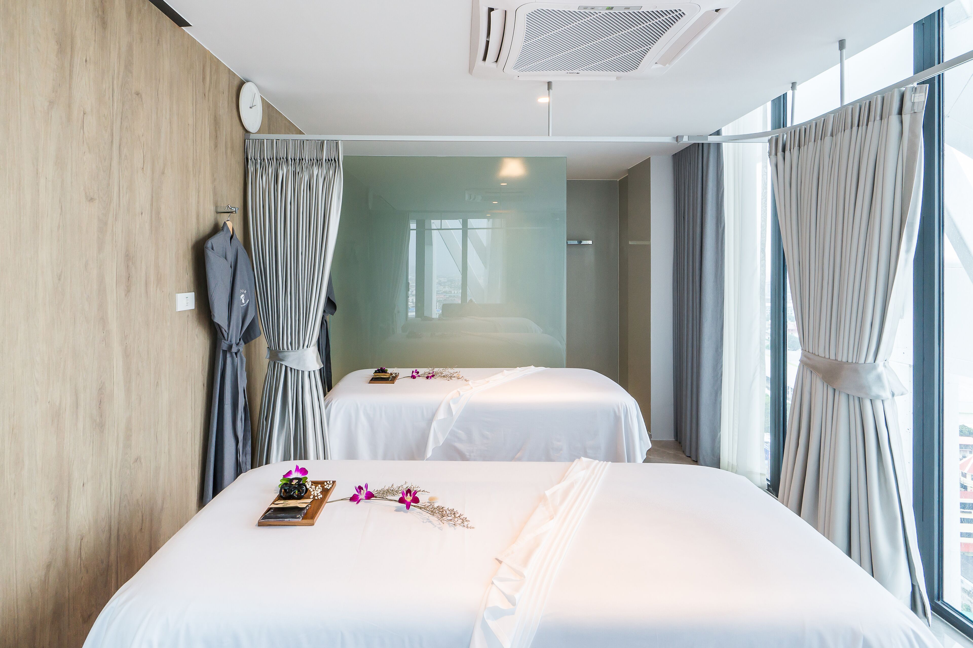 A hotel room with two beds, a wooden wall, a clock, and a large window with curtains.