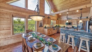 A wooden cabin with a dining area set for six, a kitchen with modern appliances, and a large window overlooking greenery. - Whiskey Mountain Lodge * Mountain View!! *Private Mountain Setting *Hot Tub *Pool Table *Ping Pong (Sevierville)