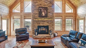 A spacious living room with a stone fireplace, leather sofas, and large windows offering views of the outdoors. - Whiskey Mountain Lodge * Mountain View!! *Private Mountain Setting *Hot Tub *Pool Table *Ping Pong (Sevierville)