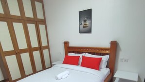 Een net opgemaakt bed met witte lakens en twee rode kussens, een houten hoofdbord, een klein nachtkastje en een aan de muur hangend schilderij.
