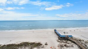 Un hôtel situé en bord de mer, avec un long bâtiment qui s’avance dans l’eau, une plage de sable et un ciel dégagé.
