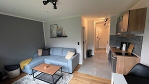 A modern living room with a sectional sofa, a coffee table, and a small rug. A kitchen with a countertop and cabinets is visible in the background. - Modern vacation apartment with terrace in Büsum (Büsum)