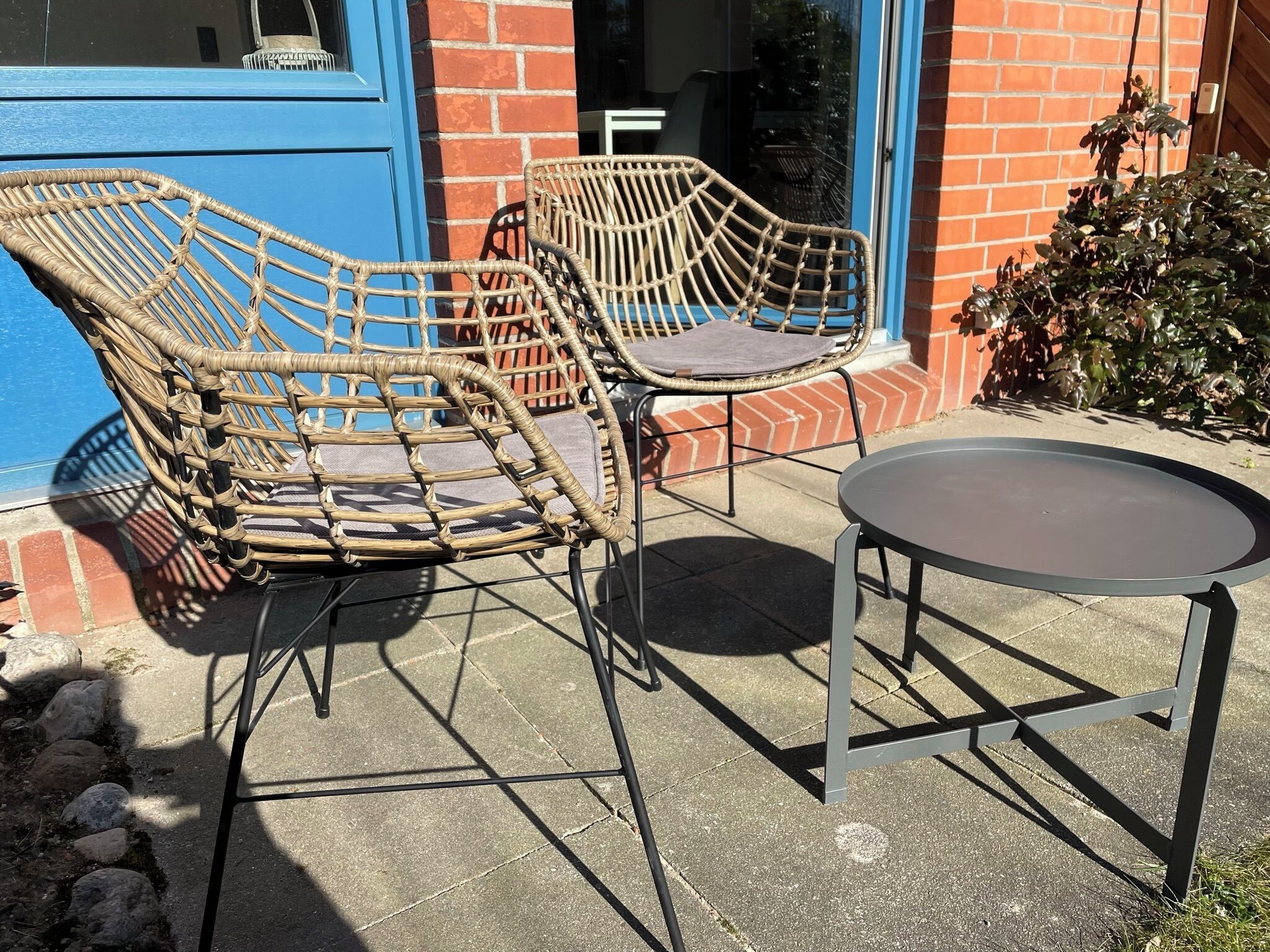 Two wicker chairs and a small round table outside a building with a blue door and brick walls.