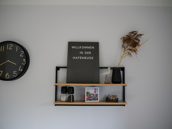 A wall clock, a welcome sign, a shelf with decorative items, and a vase with dried plants.