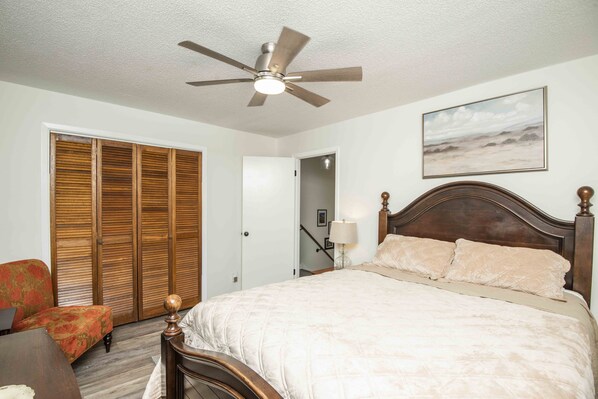 A bedroom with a bed, a ceiling fan, a wooden headboard, a nightstand, a lamp, a chair, and a closet with sliding doors.