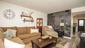 A living room with a brown sofa, a coffee table, a clock, shelves with decorative items, and a cityscape mural. - A Taste of Tennessee (Knoxville, TN)