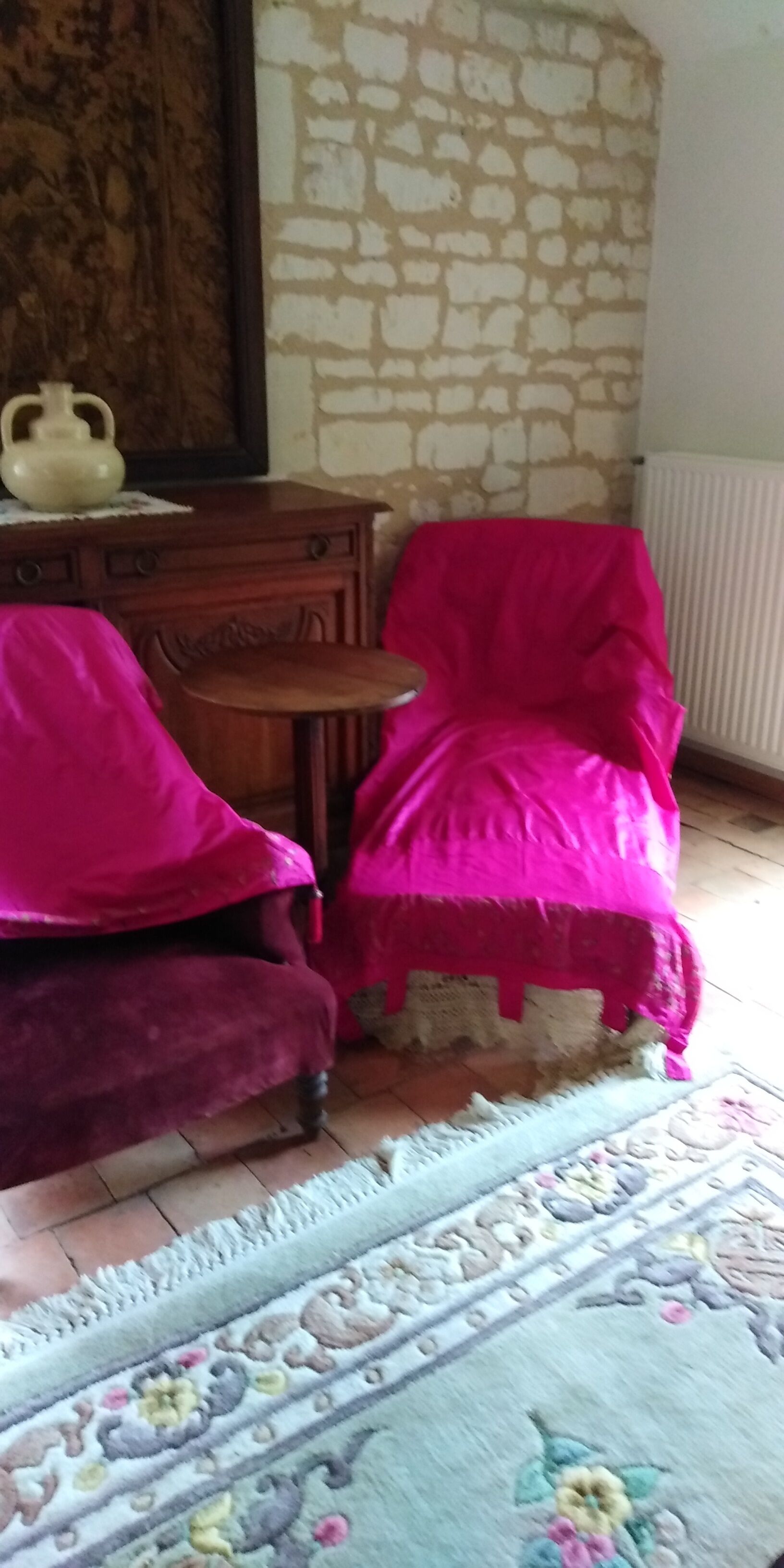 A room with a stone wall, a wooden cabinet, a small table, and two chairs covered in pink fabric.