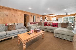 A cozy living room with a wooden wall, a large sofa, a smaller sofa, a coffee table, and a kitchen in the background.