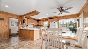 A spacious kitchen with wooden cabinets, a central island, and a dining area with white chairs. - HOT TUB! GAME ROOM w/LEDs! CLOSE TO EVERYTHING! EV Charger! Adorable décor. (Big Bear City)
