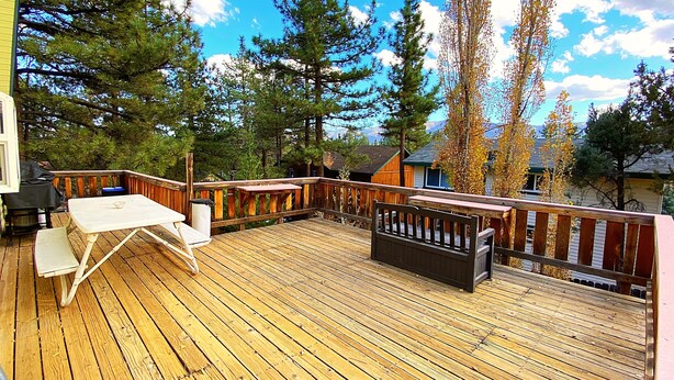 A wooden deck with a table and chairs, surrounded by tall trees and a small cabin. - HOT TUB! GAME ROOM w/LEDs! CLOSE TO EVERYTHING! EV Charger! Adorable décor. (Big Bear City)