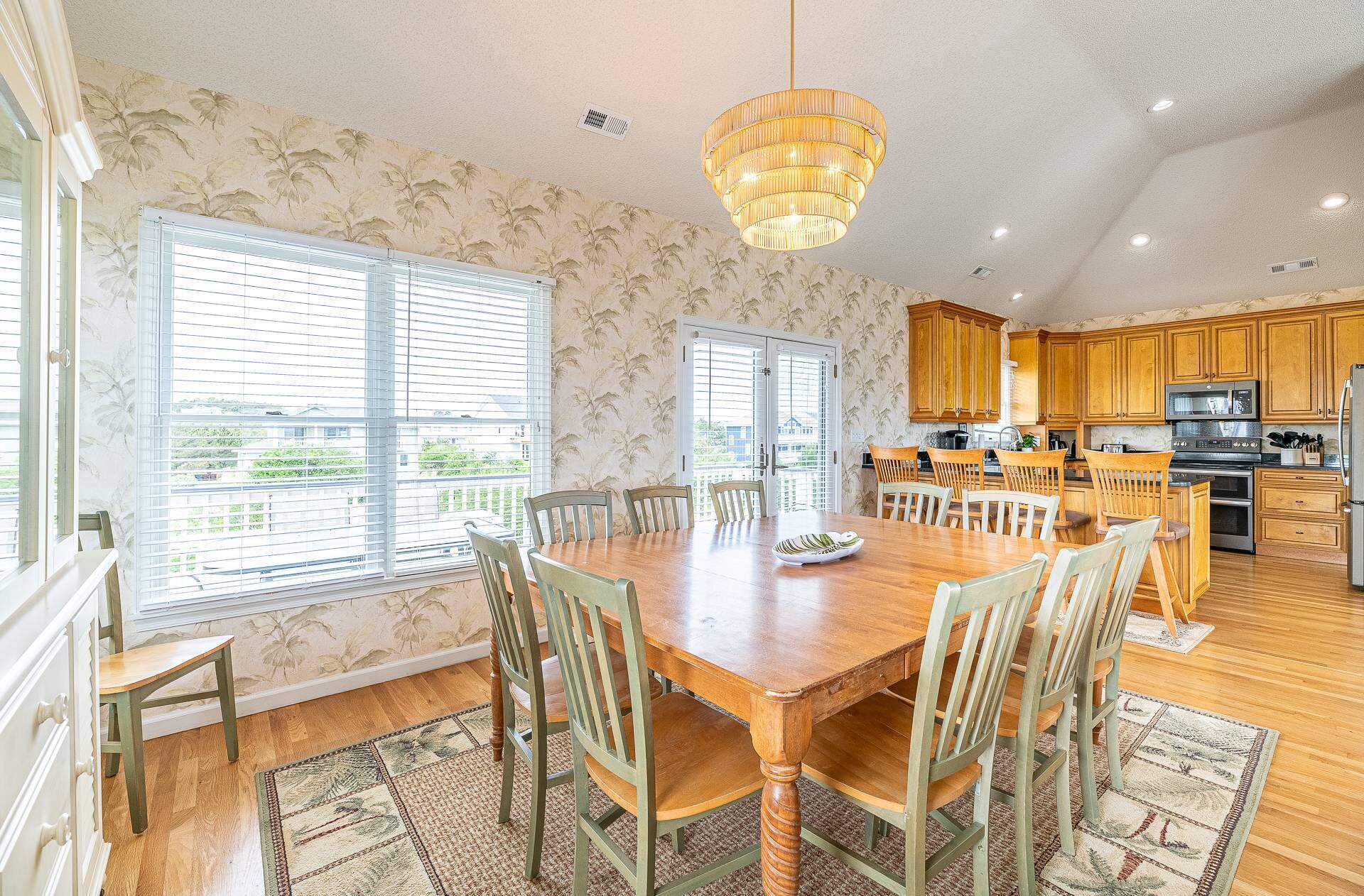 A spacious dining area with a wooden table and chairs, a large chandelier, and a view through the windows.
