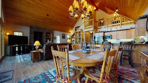 Un vaste salon doté d’une cheminée, d’une table à manger en bois et de sièges confortables. On aperçoit au fond une cuisine avec des meubles en bois et un coin repas.