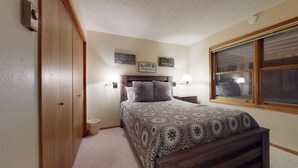 A bedroom with a wooden bed, a nightstand, a wall-mounted lamp, and a window with blinds.