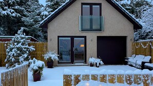 Uma casa de dois andares com garagem, cercada por um muro de madeira e árvores cobertas de neve.