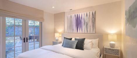 Un dormitorio con una cama grande, dos mesitas de noche con lámparas, un ventanal corredero a un patio, y un cuadro abstracto moderno en la pared.