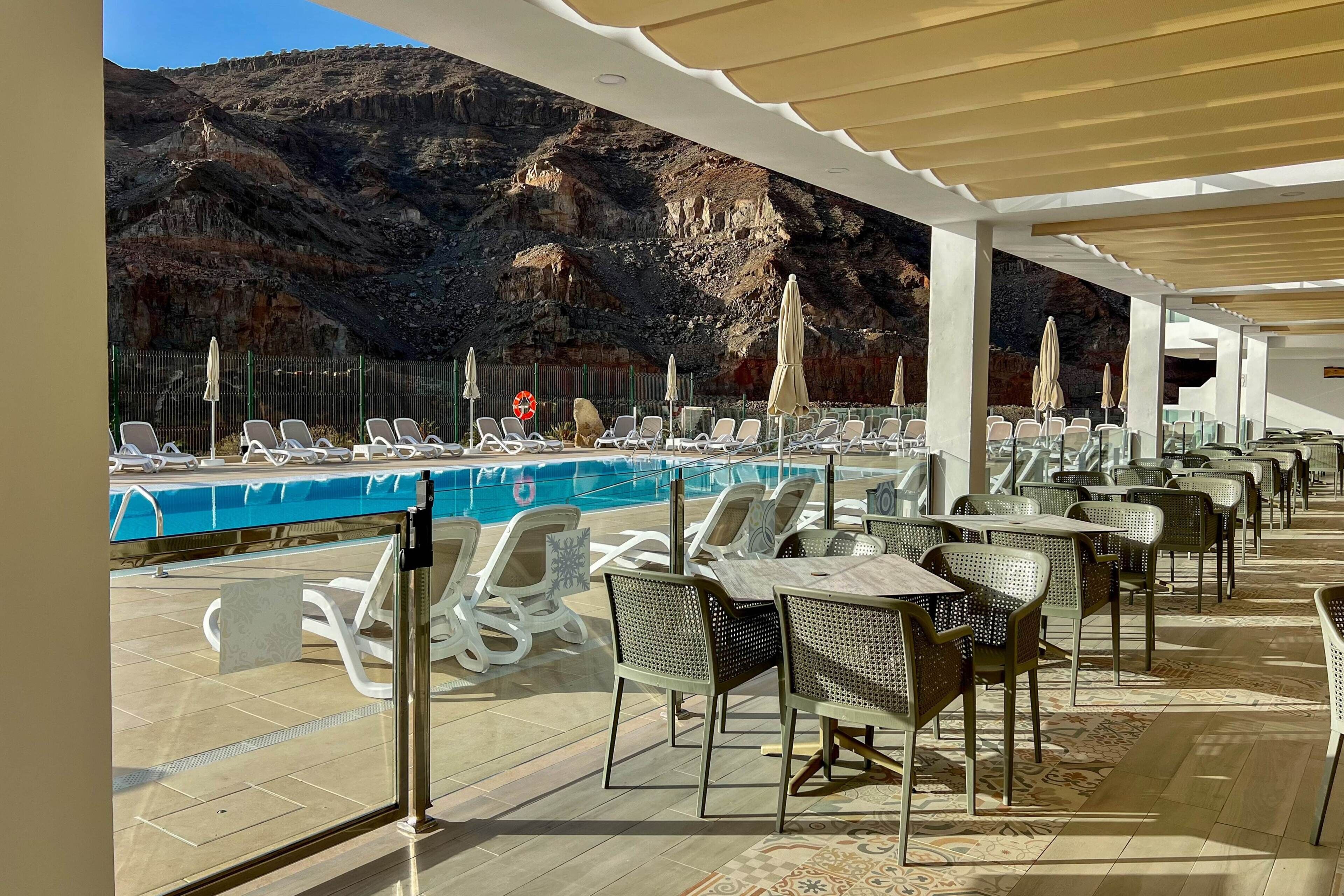 A poolside area with lounge chairs and tables, a pool, and a mountainous backdrop.