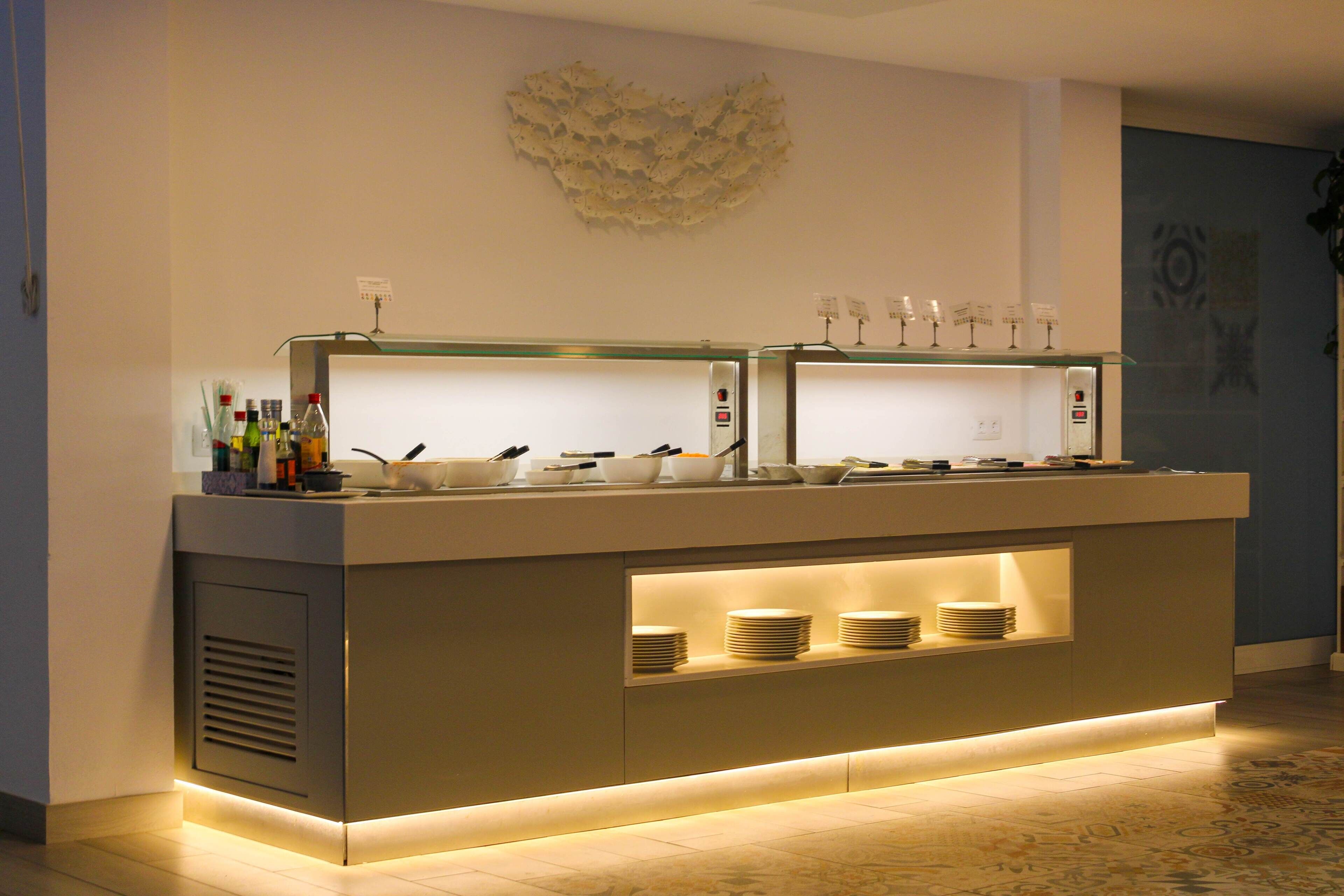 A modern buffet counter with a glass top, a decorative wall piece above, and a built-in shelf with stacked plates.