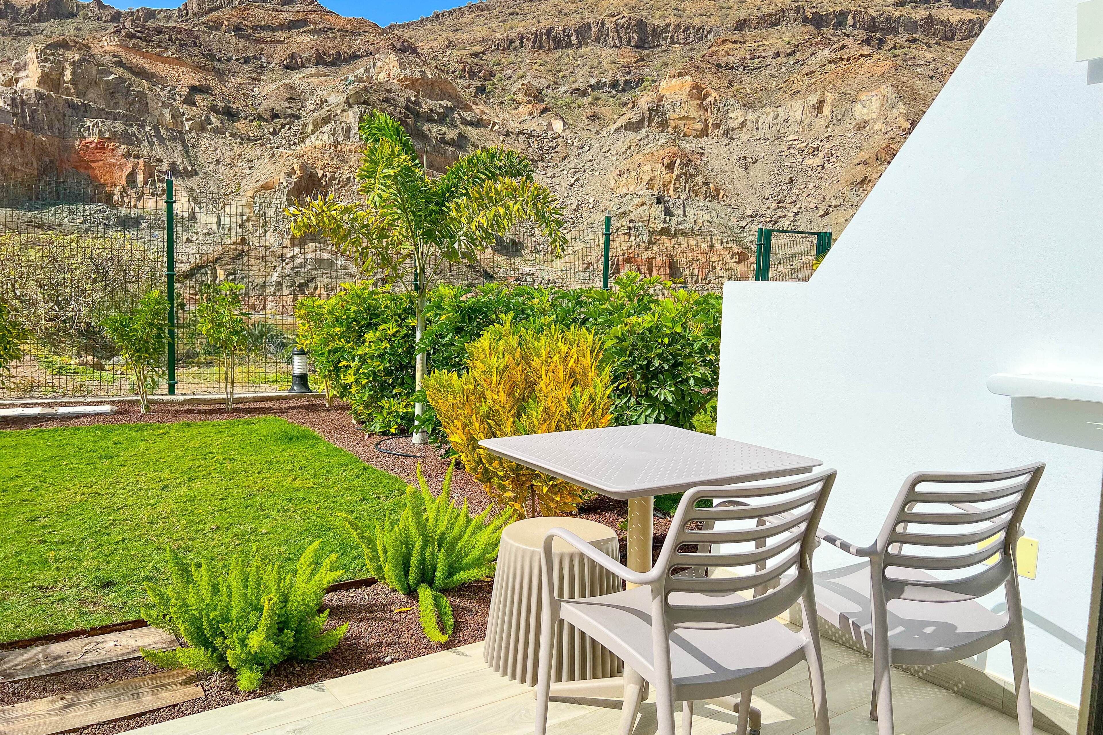 Een terras met een tafel en stoelen, omringd door een tuin met diverse planten en uitzicht op een rotsachtig landschap.