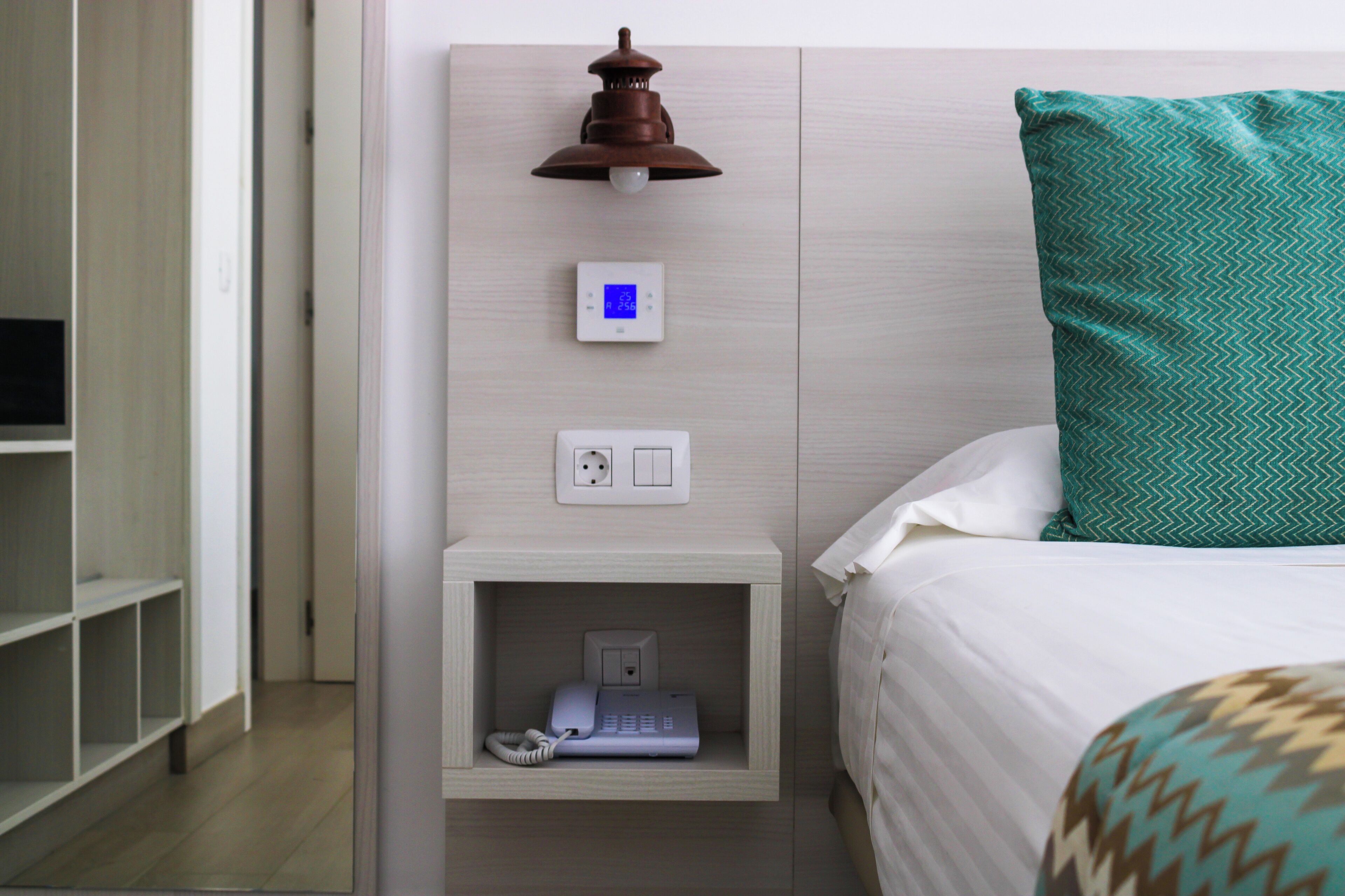 Habitación de hotel con cama, mesita de noche con lámpara, termostato y teléfono.