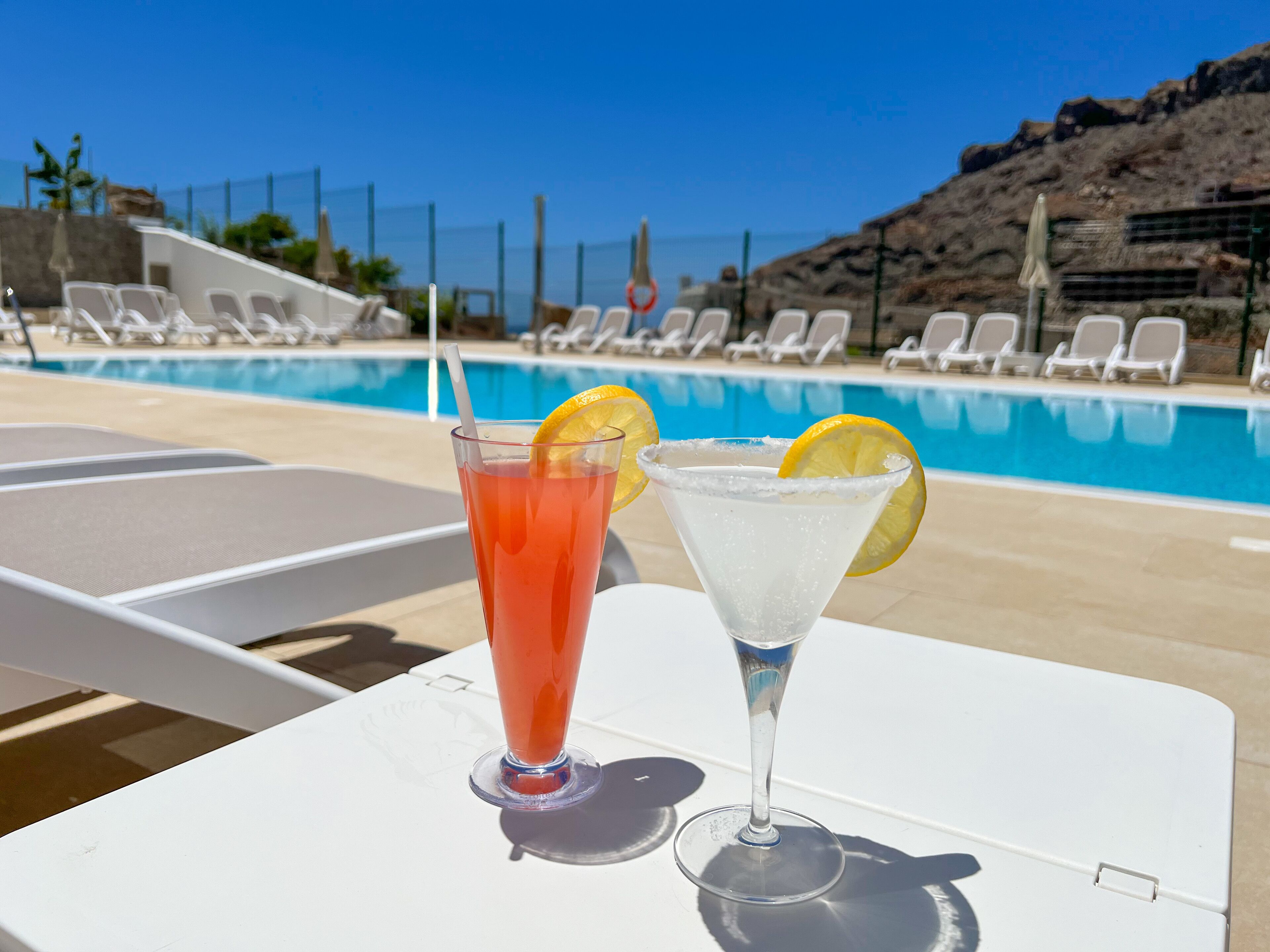 Deux boissons sur une table près d’une piscine, avec des chaises longues et une colline rocheuse en arrière-plan.