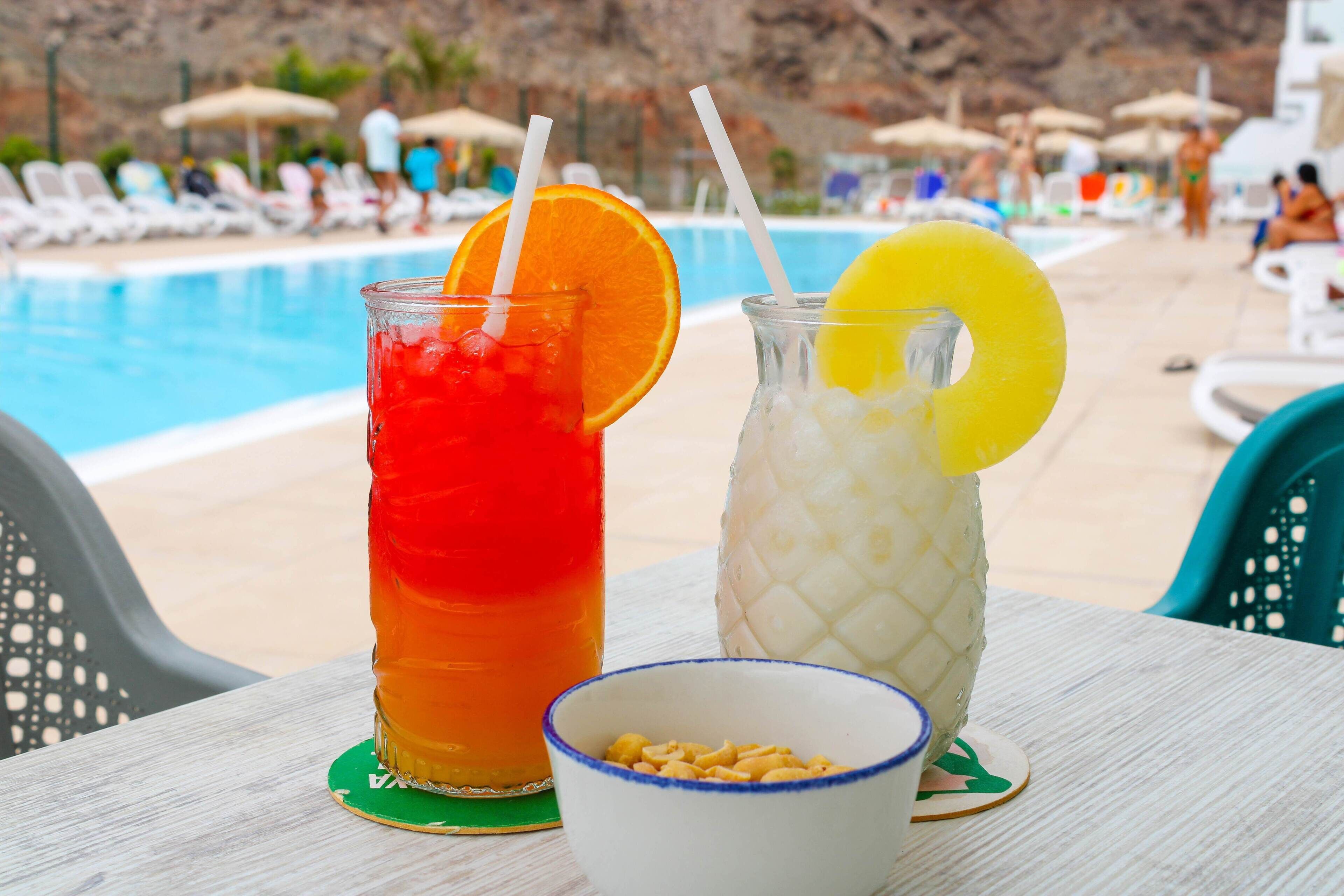 Dos bebidas con rodajas de fruta sobre una mesa junto a una piscina, con sillones y sombrillas al fondo.