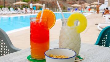 Dos bebidas con rodajas de fruta sobre una mesa junto a una piscina, con sillones y sombrillas al fondo.