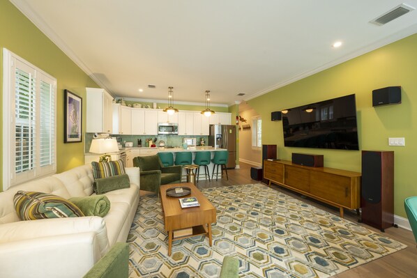 A modern living room with a sofa, coffee table, and patterned rug. A kitchen with bar stools and a TV is visible in the background.